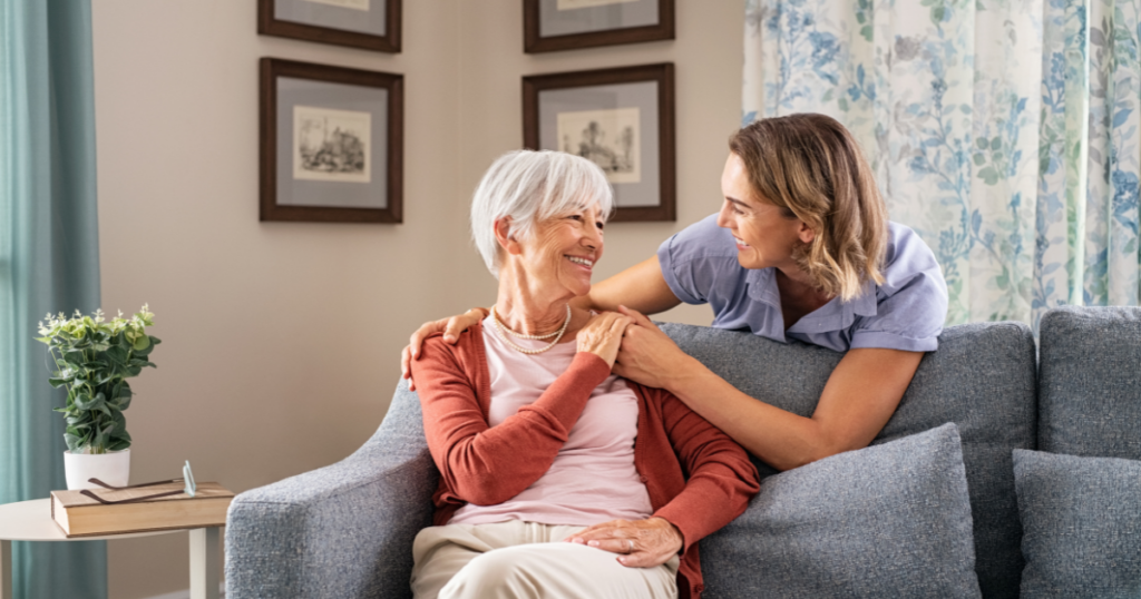 Smiling caregiver with hand over the shoulder of a happy elderly woman sitting on the couch, representing the results of quality home care services.